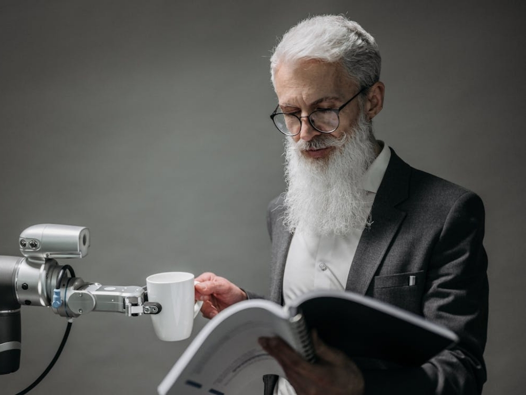 Bearded man reading a spiral bound book while a robot holds his coffee cup